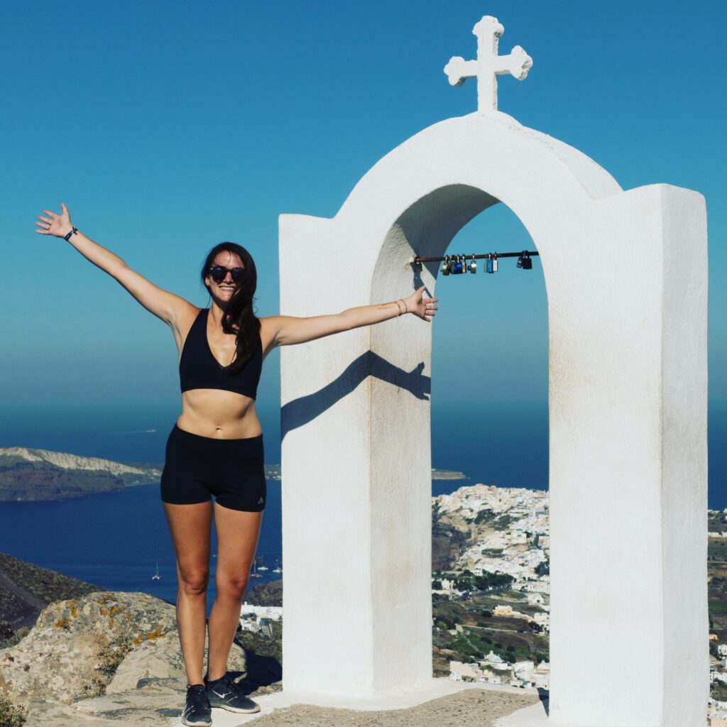 Cortney in a black workout outfit in Santorini on a hike by a white structure with a cross